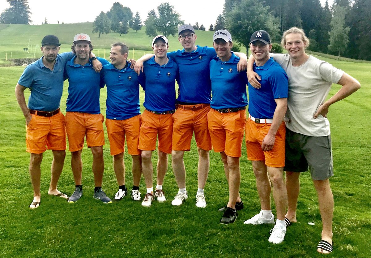 Golfteam-Gruppenfoto auf dem Golfplatz: acht Männer in blauen Poloshirts und orangen Shorts, Arm in Arm.