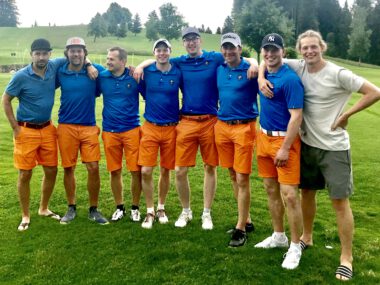 Golfteam-Gruppenfoto auf dem Golfplatz: acht Männer in blauen Poloshirts und orangen Shorts, Arm in Arm.