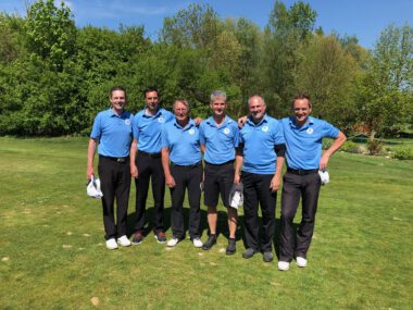 Golfteam im Mannschaftsfoto auf dem Golfplatz – sechs Spieler in blauen Poloshirts posieren bei Sonne vor grüner Kulisse