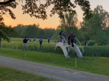 Golfer beim Putten auf dem Green bei Sonnenuntergang, Zuschauer auf Bogenbrücke am Teich auf dem Golfplatz