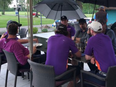 Golfer-Team in lila Shirts entspannt auf Clubhaus-Terrasse unter Schirmen, Drinks auf dem Tisch, Golfplatz im Hintergrund
