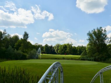Sommerlicher Golfplatz: gepflegtes Green mit Fahne, Teich mit Fontäne und Brücke, umgeben von Bäumen und blauem Himmel.