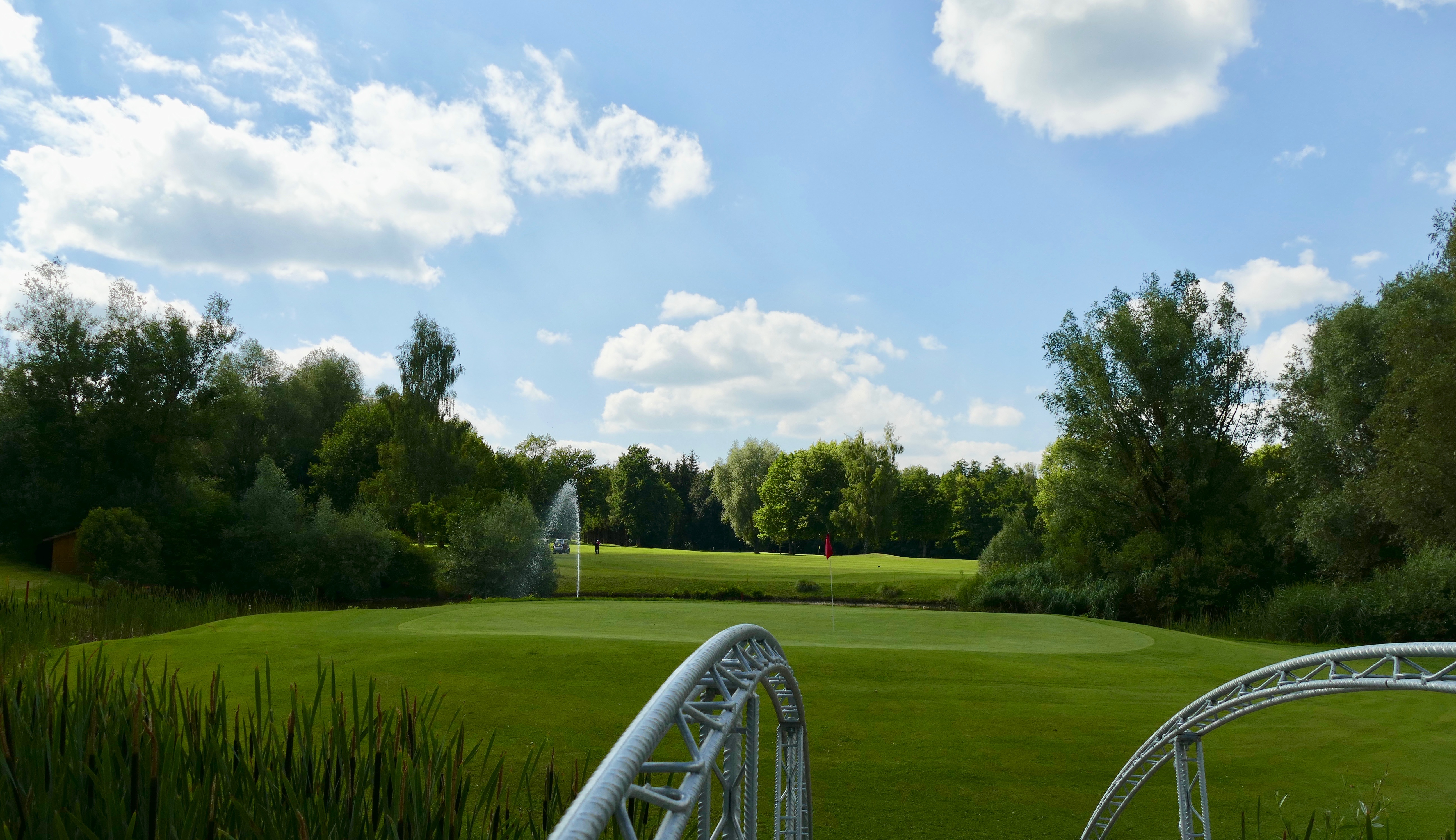 Sommerlicher Golfplatz: gepflegtes Green mit Fahne, Teich mit Fontäne und Brücke, umgeben von Bäumen und blauem Himmel.