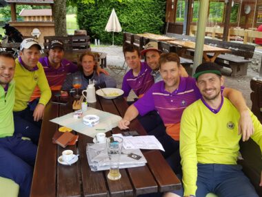 Golfteam entspannt nach Turnier im Clubhaus: lachende Gruppe in bunten Trikots beim Essen auf der Terrasse.