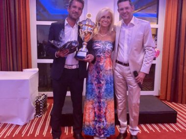 Preisverleihung: Drei Personen posieren mit großer Trophäe auf Bühne bei Gala-Event; Siegerfoto mit Pokal und Moderator.