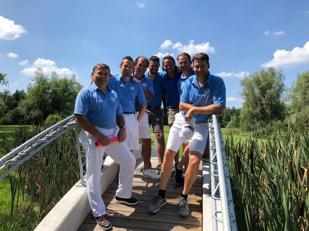 Golfclub-Team in blauen Poloshirts auf Brücke im Grünen – fröhliches Gruppenfoto bei Sonnenschein