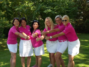 Damen-Golfteam in pinken Poloshirts und weißen Röcken und Shorts, Daumen hoch, Gruppenfoto im Grünen auf dem Golfplatz