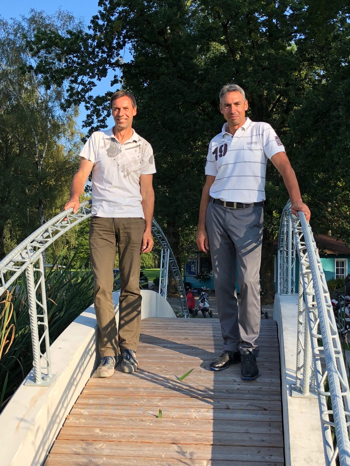 Zwei Männer stehen auf einer kleinen Fußgängerbrücke im Park bei Sonnenschein, Holzsteg mit Metallgeländer, grüne Bäume.