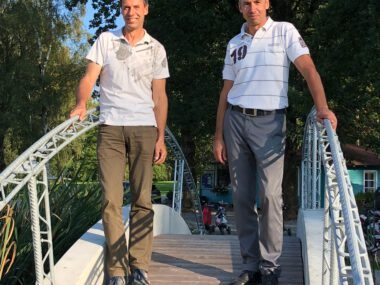 Zwei Männer stehen auf einer kleinen Fußgängerbrücke im Park bei Sonnenschein, Holzsteg mit Metallgeländer, grüne Bäume.