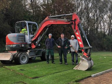 GaLaBau-Team mit rotem Bagger verlegt Rollrasen auf Sportplatz – Erdarbeiten und Landschaftsbau