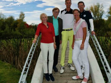 Fünf Golfer posieren lächelnd auf einer Holzbrücke im Golfclub, grüner Platz und herbstliche Bäume im Hintergrund