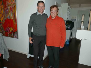 Zwei Männer lächeln in moderner Wohnküche; einer hält Mikrofon, Kunstbild im Hintergrund, Porträtfoto indoor