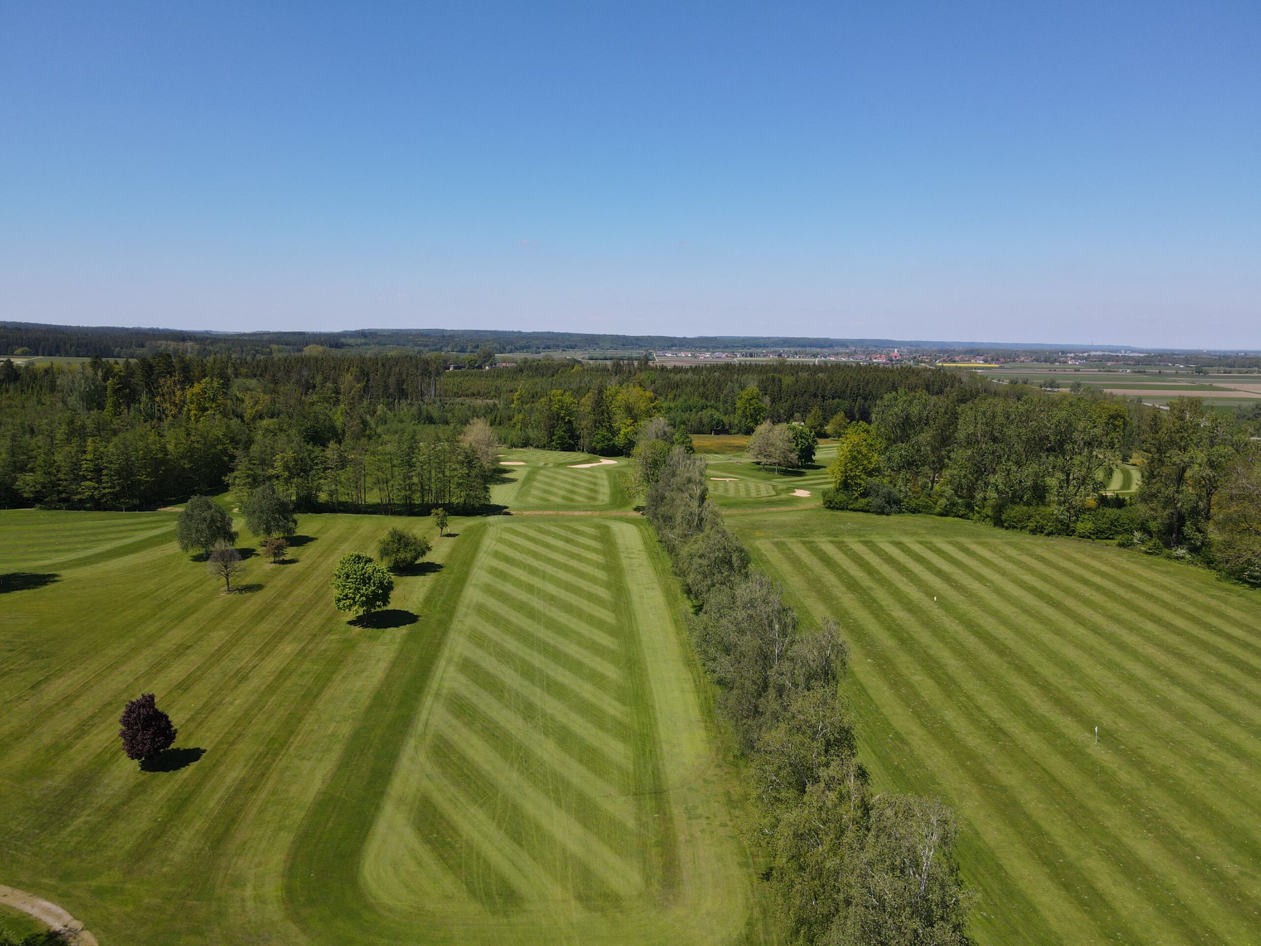 Golfplatz Luftaufnahme: gestreifte Fairways, Baumreihen und Sandbunker in grüner Landschaft unter blauem Himmel