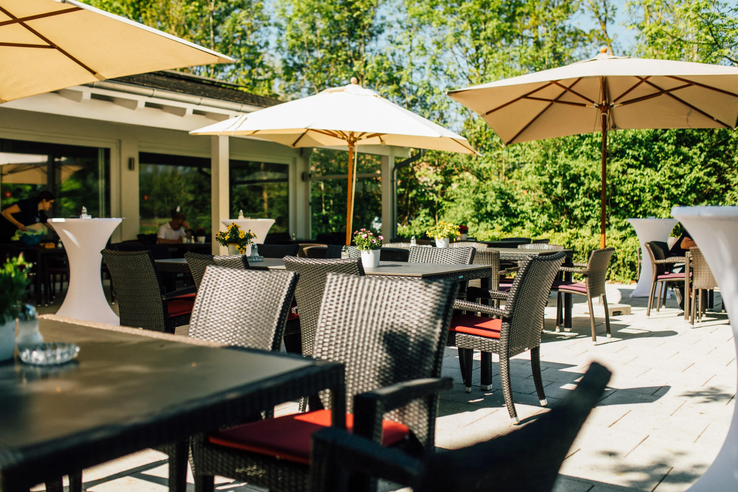 Restaurant-Terrasse im Grünen mit Rattanmöbeln, Sonnenschirmen und Blumen – gemütlicher Außenbereich zum Essen im Sommer
