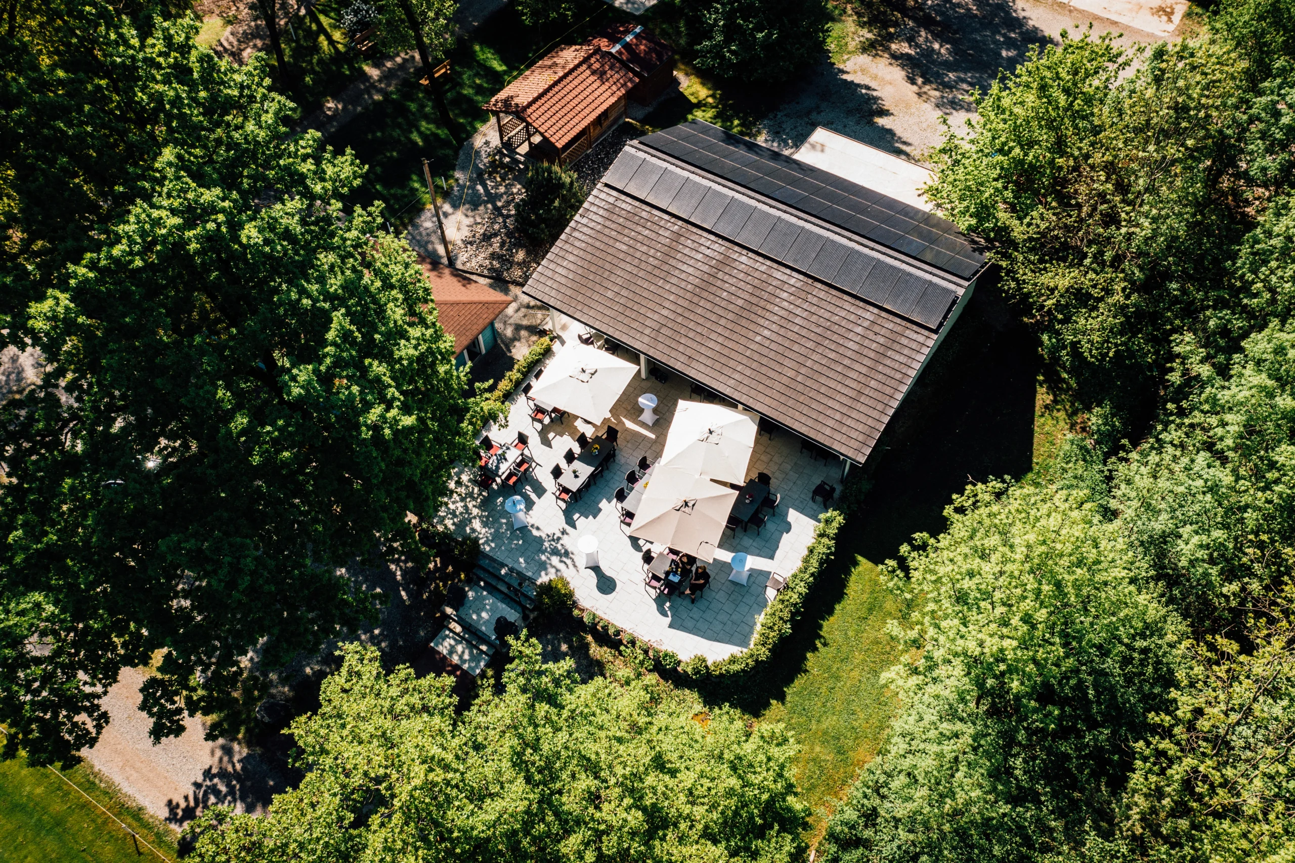 Luftaufnahme Restaurant-Biergarten im Grünen mit Sonnenschirmen, Gästen und Gebäude mit Solardach