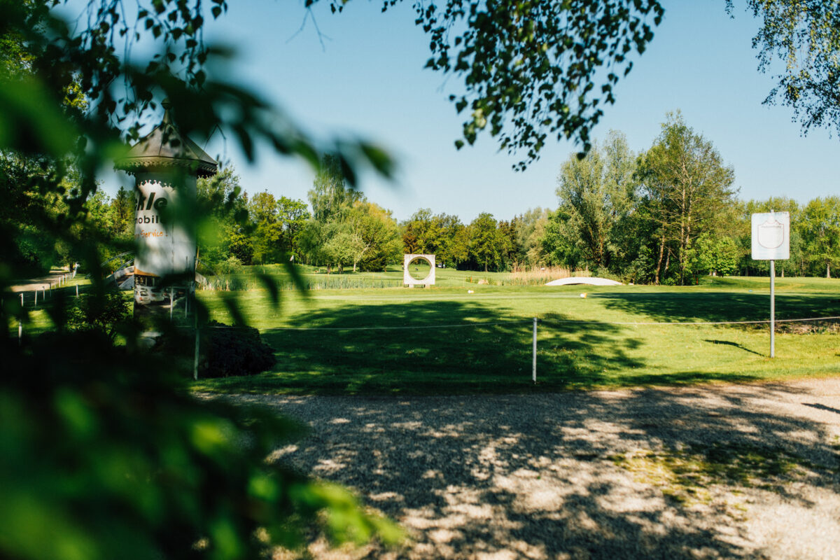 Golfplatz in Parklandschaft bei Sonnenschein mit Teich, kleiner Brücke und runder Steinskulptur – ruhige Naturidylle
