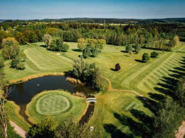 Luftaufnahme Golfplatz mit Green, Fairways, Teich und Brücke in grüner Landschaft
