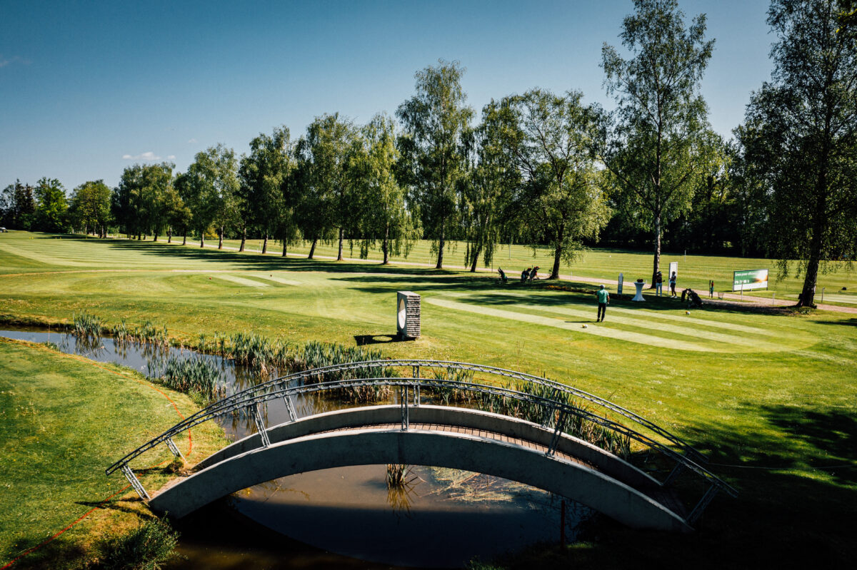 Sonniger Golfplatz mit Brücke über Teich, Golfer am Abschlag auf gepflegtem Fairway zwischen Birken.